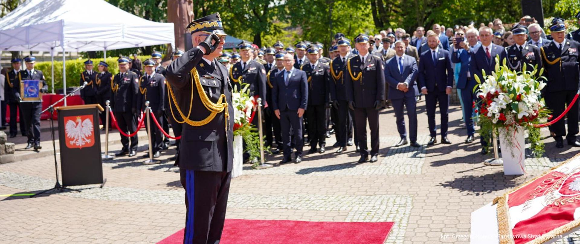 Zdjęcie 3 a.jpeg stanowi kolejny kadr z tej samej uroczystości państwowej, ukazując moment składania meldunku lub oddawania honorów w kluczowym punkcie ceremonii.
Oto szczegółowy opis elementów widocznych na fotografii:
Główny plan
Salutujący oficer: Na pierwszym planie, lekko po lewej stronie, stoi wysoki rangą oficer Państwowej Straży Pożarnej w pełnym umundurowaniu galowym (czarna czapka garnizonowa ze złotymi zdobieniami, złote sznury galowe, ciemny mundur z niebieskimi lampasami na spodniach). Oficer stoi na baczność na czerwonym dywanie i salutuje w stronę prawej krawędzi kadru.
Sztandar: W prawym dolnym rogu widoczny jest fragment ułożonego na postumencie nowego sztandaru o barwach narodowych, przed którym oficer prawdopodobnie oddaje honory.
Tło i pozostali uczestnicy
Szyk mundurowy i zaproszeni goście: Za salutującym oficerem, w głębi placu, stoi rządem liczna grupa osób. Są wśród nich pozostali funkcjonariusze straży pożarnej w galowych mundurach oraz cywile w garniturach (w tym reprezentanci władz i instytucji partnerskich).
Sceneria: Po lewej stronie kadru widoczna jest mównica z godłem Polski (orłem w koronie) oraz biały namiot ogrodowy. W tle wznosi się ciemny pomnik, a całe otoczenie otaczają gęste, zielone korony drzew w pełnym letnim słońcu. Po prawej stronie i w centrum ustawiono eleganckie kompozycje z białych i czerwonych kwiatów.
W prawym dolnym rogu fotografii znajduje się podpis autora zdjęcia: kpt. Grzegorz Nowak | Państwowa Straż Pożarna.