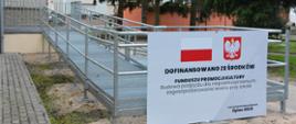 Metalowa rampa z poręczami prowadząca do wejścia budynku; na pierwszym planie tablica informacyjna z polską flagą i godłem, informująca o dofinansowaniu budowy podjazdu dla osób z niepełnosprawnościami ze środków Funduszu Promocji Kultury.