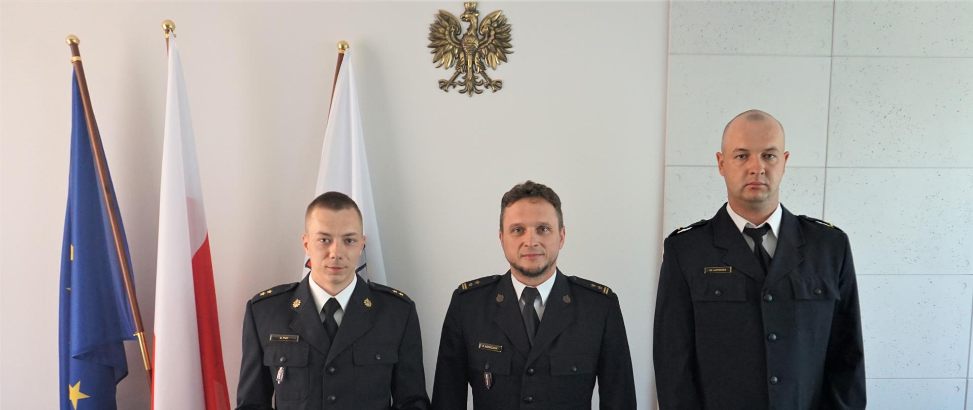 Na tle białej ściany stoi trzech strażaków w granatowych mundurach. U góry złoty orzeł. Po lewej flagi Polski UE i godło PSP.