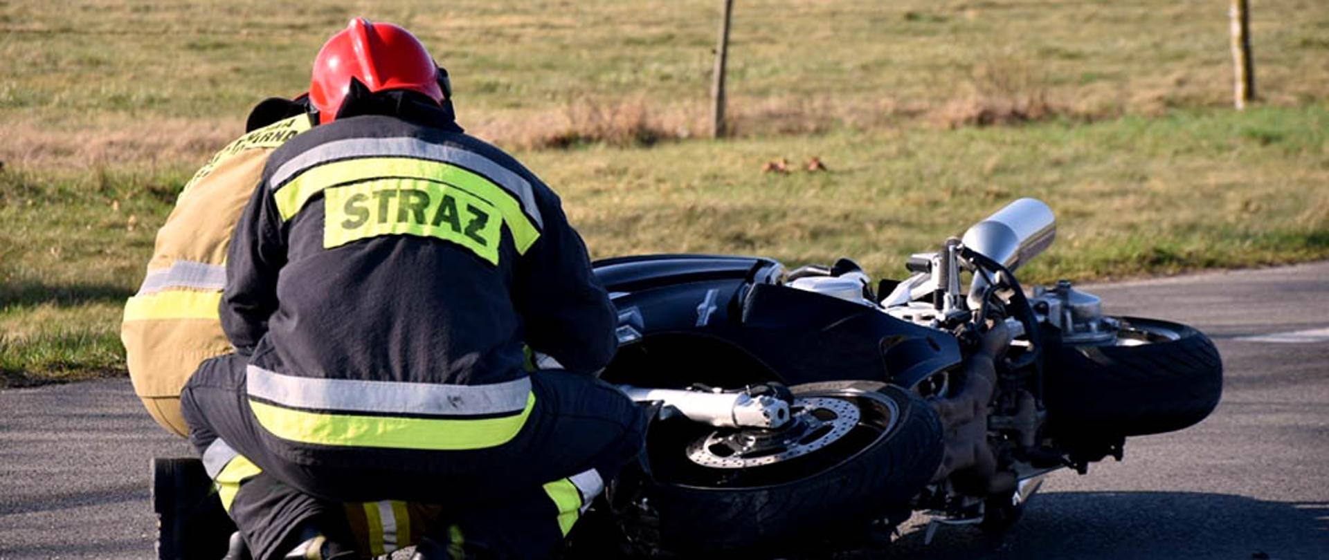 Na zdjęciu dwóch strażaków zabezpiecza przewrócony na wskutek wypadku motocykl który leży na jezdni.