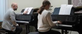 zdjęcie przedstawia uczennicę grającą na fortepianie w sali kameralnej, w tle prowadzący warsztaty przy drugim fortepianie