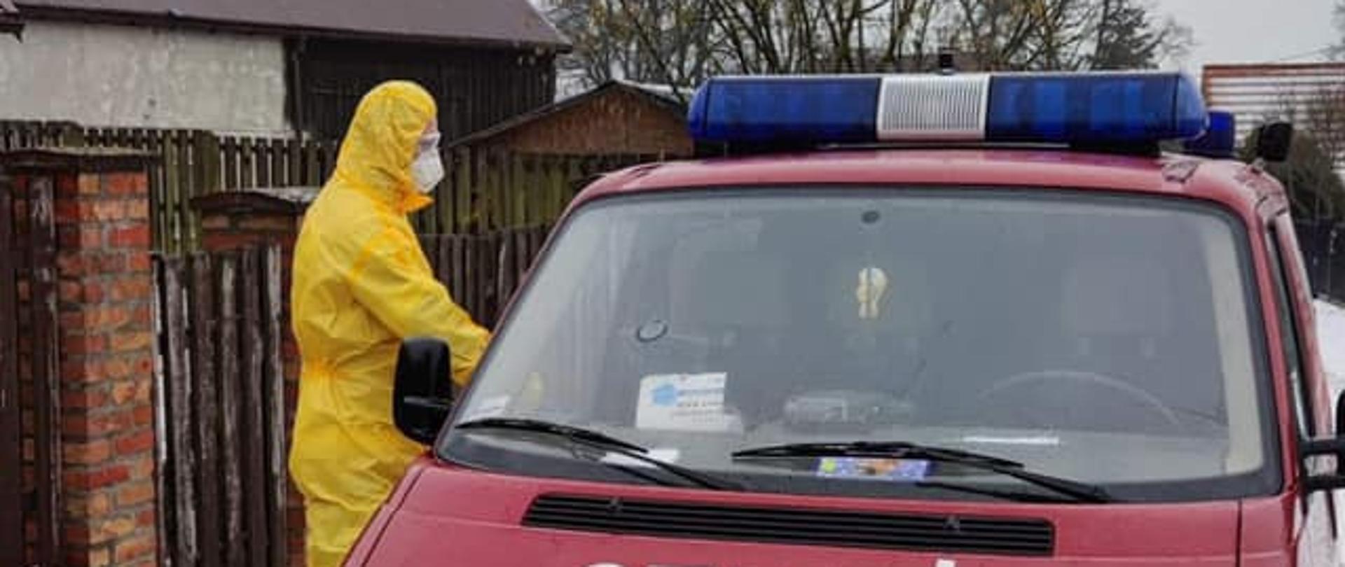 Na zdjęciu znajduje się wóz strażacki. Po lewej stronie wozu strażackiego znajduje się strażak ubrany w żółty kombinezon ochronny. 