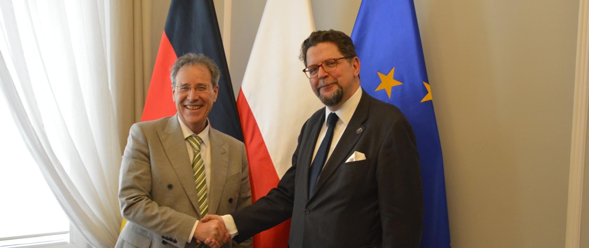 Wiceminister Szeptycki stoi obok mężczyzny w jasnoszarym garniturze, podają sobie ręce, za nimi pod żółtą ścianą flagi Polski, Niemiec i UE, z lewej przysłonięte firanką okno.