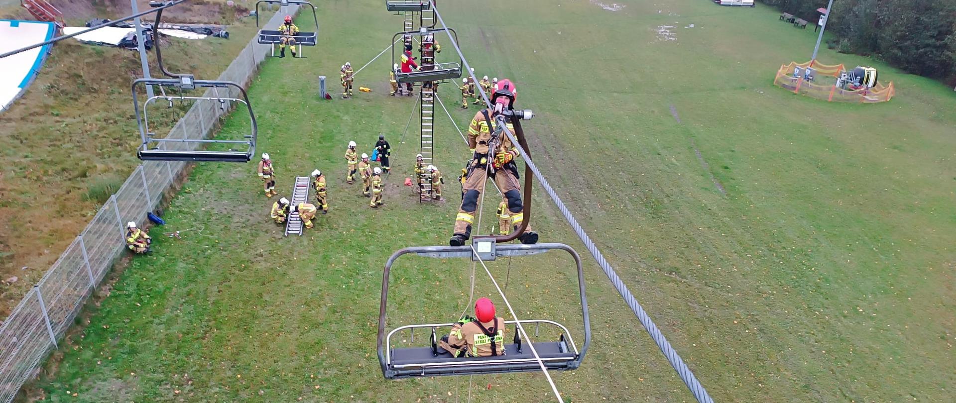 Widok z góry na ćwiczenia ratownicze strażaków przy wyciągu krzesełkowym. Jedna osoba znajduje się na krześle, druga na drabinie, a pozostali na ziemi obsługują liny i sprzęt. Teren zielony, ogrodzony, w tle budynki i sprzęt treningowy.