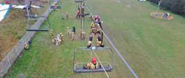 Widok z góry na ćwiczenia ratownicze strażaków przy wyciągu krzesełkowym. Jedna osoba znajduje się na krześle, druga na drabinie, a pozostali na ziemi obsługują liny i sprzęt. Teren zielony, ogrodzony, w tle budynki i sprzęt treningowy.