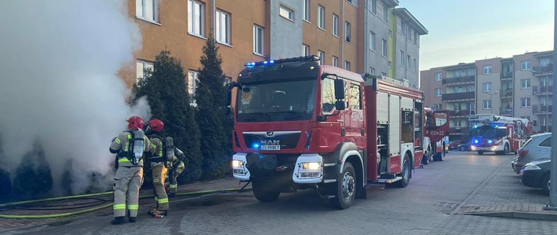 Strażacy stojący przed klatką schodową, w tle auto strażackie