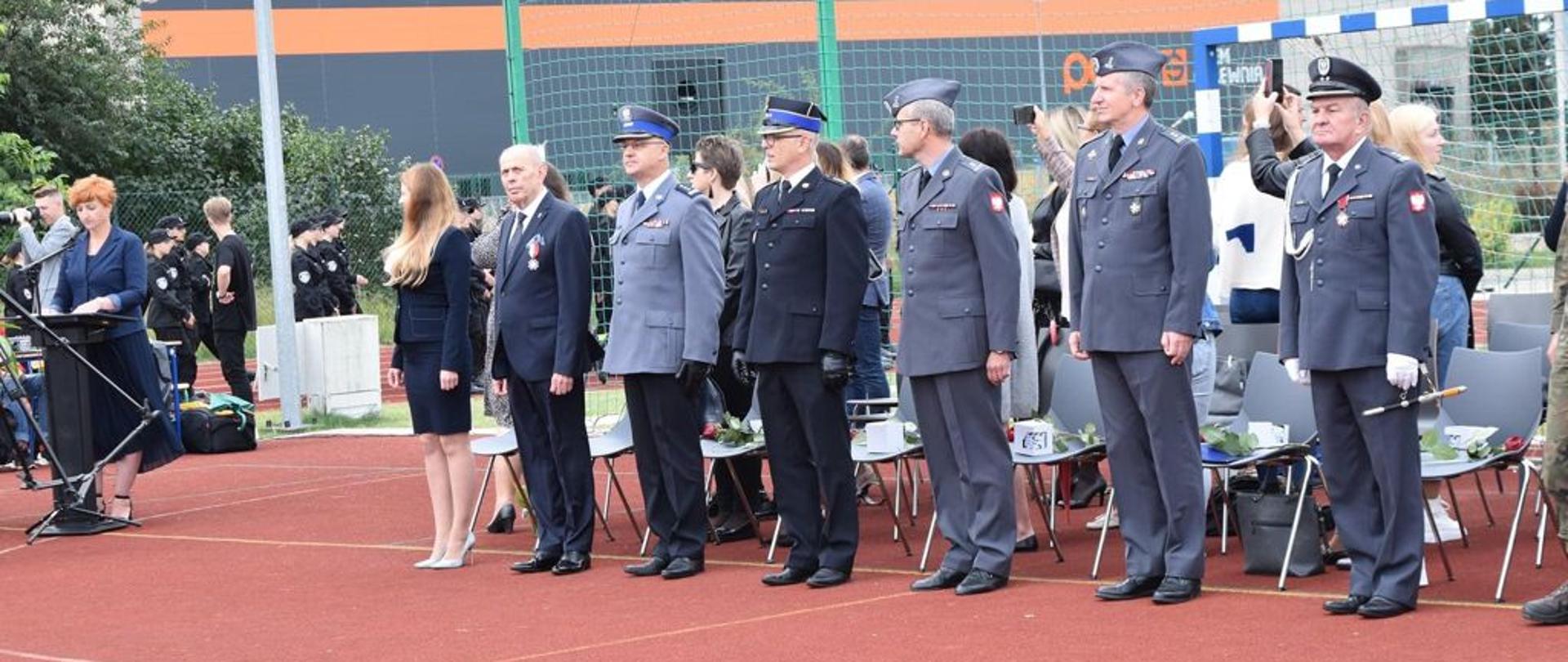 Na zdjęciu dyrektor szkoły oraz zaproszeni goście za nimi rodzice uczniów z lewej strony osoba prowadząca uroczysty apel