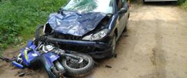 Na zdjęciu można zauważyć granatowy samochód po zderzeniu z motocyklem. Ma rozbitą przednią szybę, zniszczoną maskę. Przed zderzakiem leży zniszczony motocykl. 