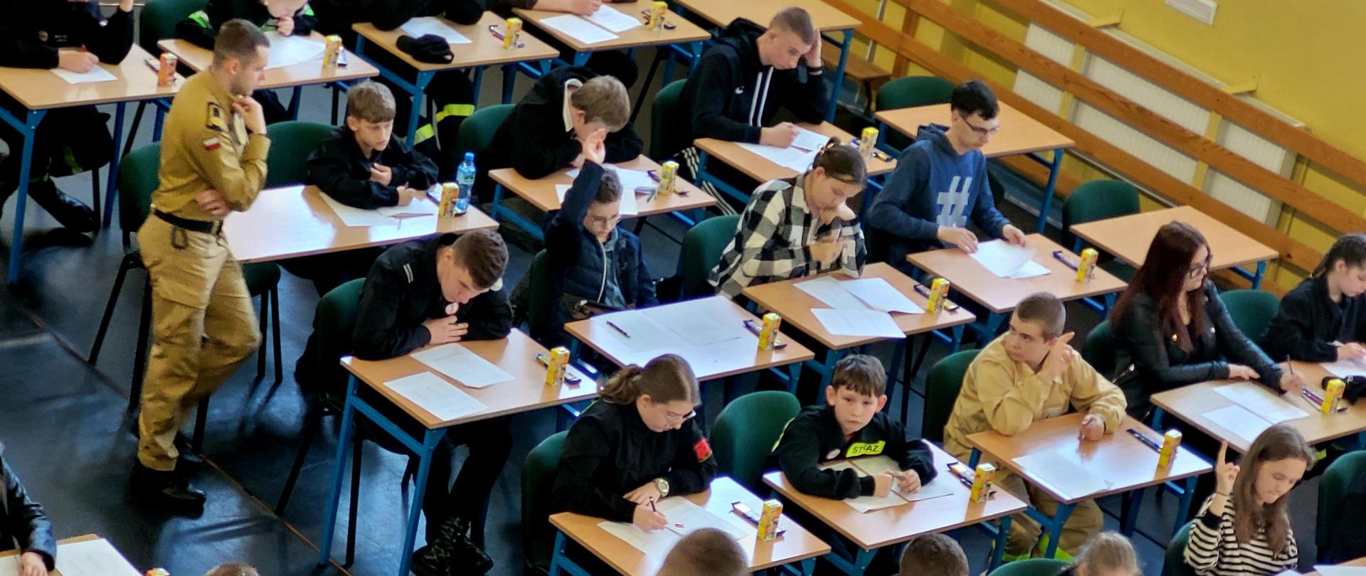 Zdjęcie z lotu ptaka. Uczestnicy siedzą przy ławkach, a członkowie komisji rozdają testy.