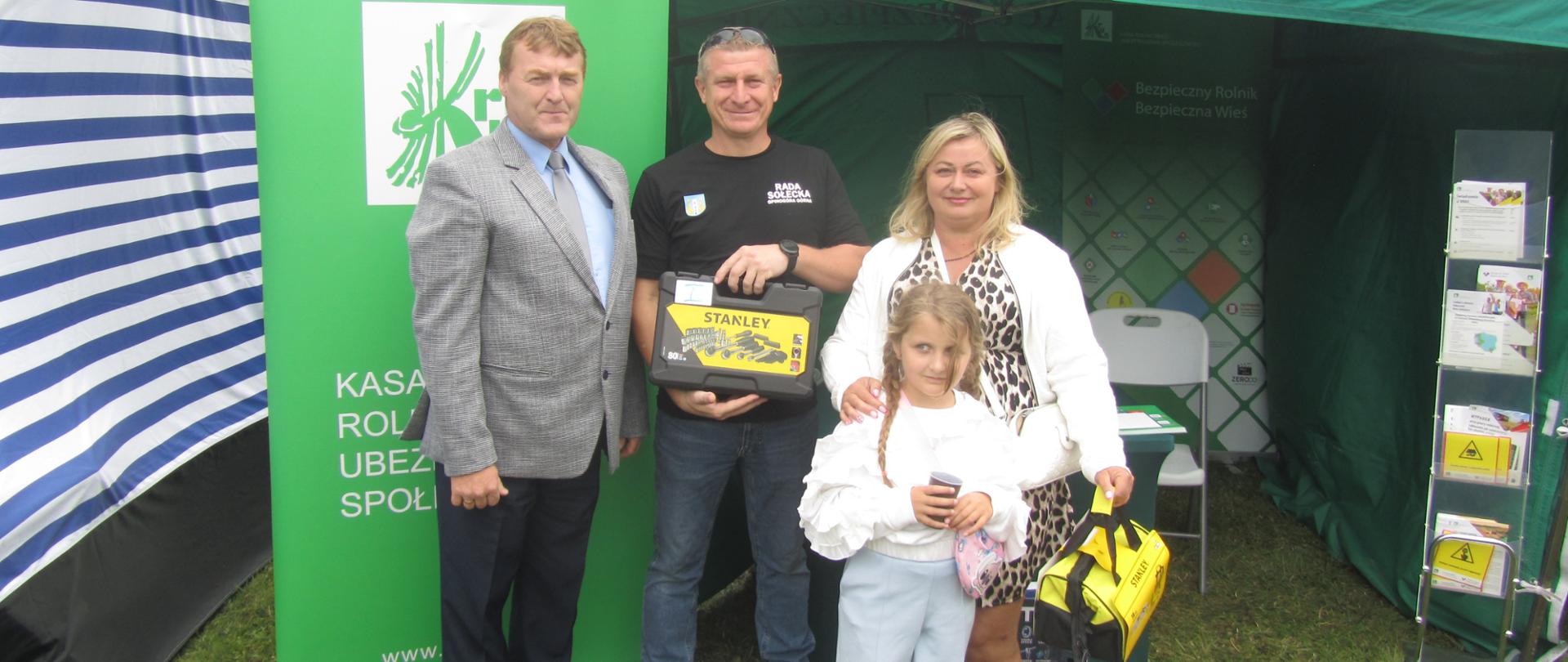 Kobieta z dziewczynką i dwóch mężczyzn pozują na tle stoiska pod zielonym namiotem KRUS. Jeden mężczyzn i kobieta trzymają w rękach skrzynki z narzędziami.