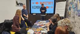 Grupa młodych ludzi siedzi wokół stołu na którym leży dużo gazet, szkicowników, wycinków, komiksów i przyborów do rysowania. na końcu sali monitor z wyświetlonym napisem "Warsztaty komiksowe", po prawej stronie gazetka ścienna.