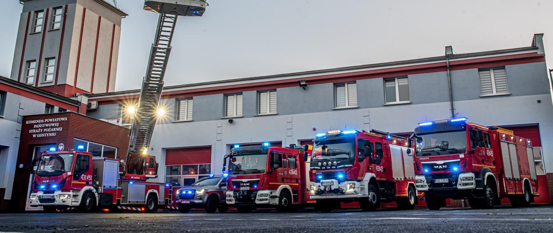  5 samochodów strażackich stoi przed budynkiem Komendy Powiatowej PSP w Gostyniu (GBA, GCBA, GBA-RT, SD oraz SLRR). Samochody mają włączone sygnały błyskowe. Kosz drabiny mechanicznej wyciągnięty jest w górę.