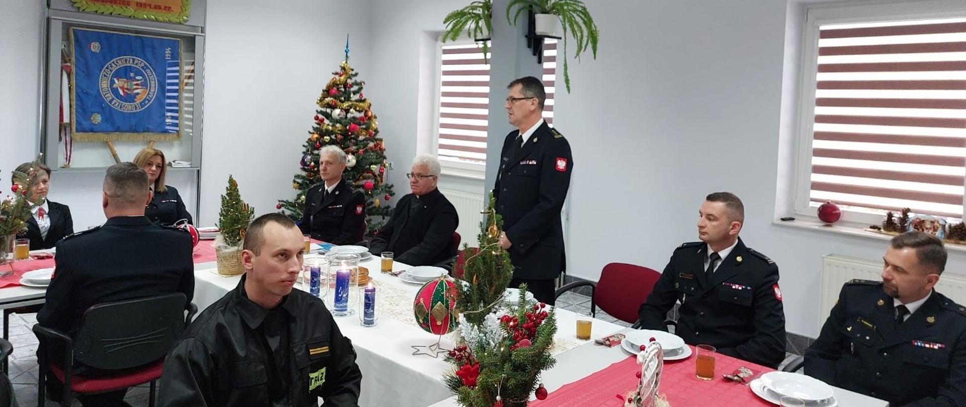 W pomieszczeniu strażacy siedzący przy udekorowanych świątecznie stołach. Jeden ze strażaków stoi i przemawia do pozostałych. W pomieszczeniu widoczna udekorowana choinka. 