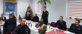 W pomieszczeniu strażacy siedzący przy udekorowanych świątecznie stołach. Jeden ze strażaków stoi i przemawia do pozostałych. W pomieszczeniu widoczna udekorowana choinka. 