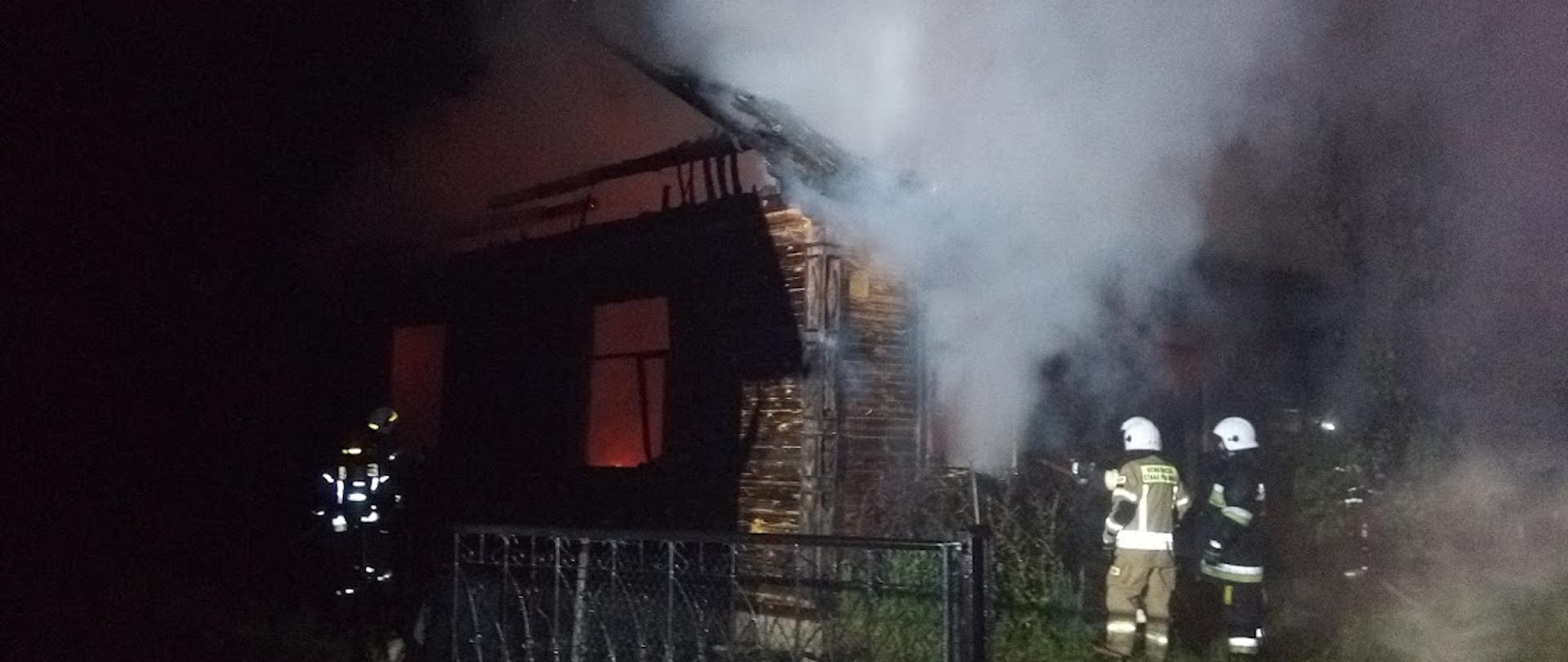 Budynek mieszkalny jednorodzinny objęty pożarem. W budynku zniszczony został dach, W szczycie budynku wypalone zostały futryny okienne. Przy budynku strażacy prowadzący działania gaśnicze. Wewnątrz budynku widoczne są płomienie. Z okien wydobywa się dym.