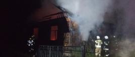 Budynek mieszkalny jednorodzinny objęty pożarem. W budynku zniszczony został dach, W szczycie budynku wypalone zostały futryny okienne. Przy budynku strażacy prowadzący działania gaśnicze. Wewnątrz budynku widoczne są płomienie. Z okien wydobywa się dym.