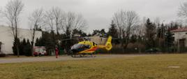 Na zdjęciu widoczny jest czarno-żółty śmigłowiec stojący na betonowym placu przy boisku szkolnym. Maszyna szykuje się do startu. Po lewej stronie widać członka załogi w czerwonym kombinezonie i czarnym hełmie na głowie. Dokonuje on inspekcji śmigłowca przed startem. W tle widać wozy strażackie z włączonymi migającymi światłami alarmowymi. Straż Pożarna zabezpiecza strat maszyny. Za helikopterem widać rząd drzew na których brak jest liści oraz budynki szkoły.