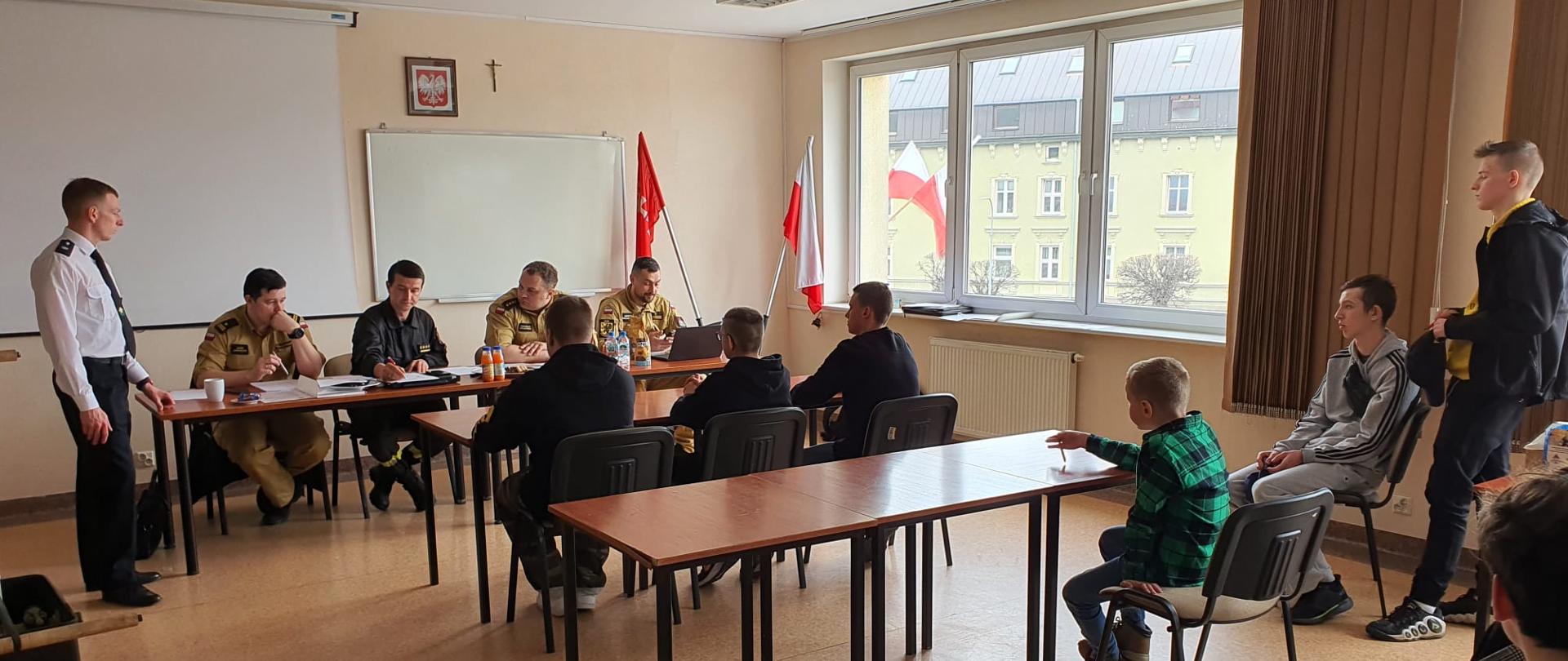 W sali szkoleniowej JRG 1 za stołem siedzi komisja KM PSP (4 strażaków), przed nimi 3 uczestników odpowiada na pytania.