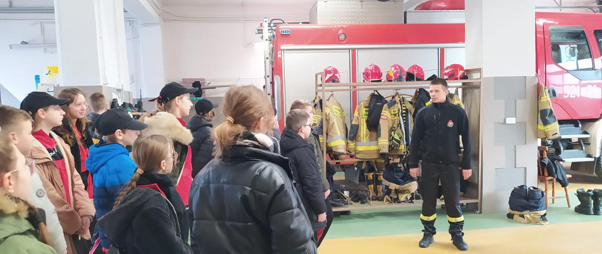 Pomieszczenie garaży jednostki ratowniczo gaśniczej w Sępólnie Krajeńskim, strażak w ubraniu koszarowym oraz kilkanaścioro dzieci w wieku szkolnym, w tle samochód ratowniczo-gaśniczy. 