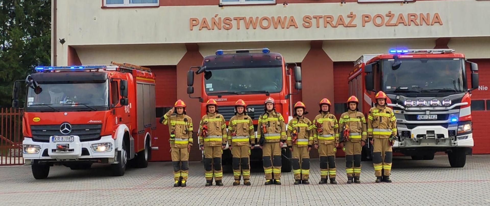 Na Zdjęciu widoczni strażacy na tle samochodów pożarniczych ora budynku JRG
