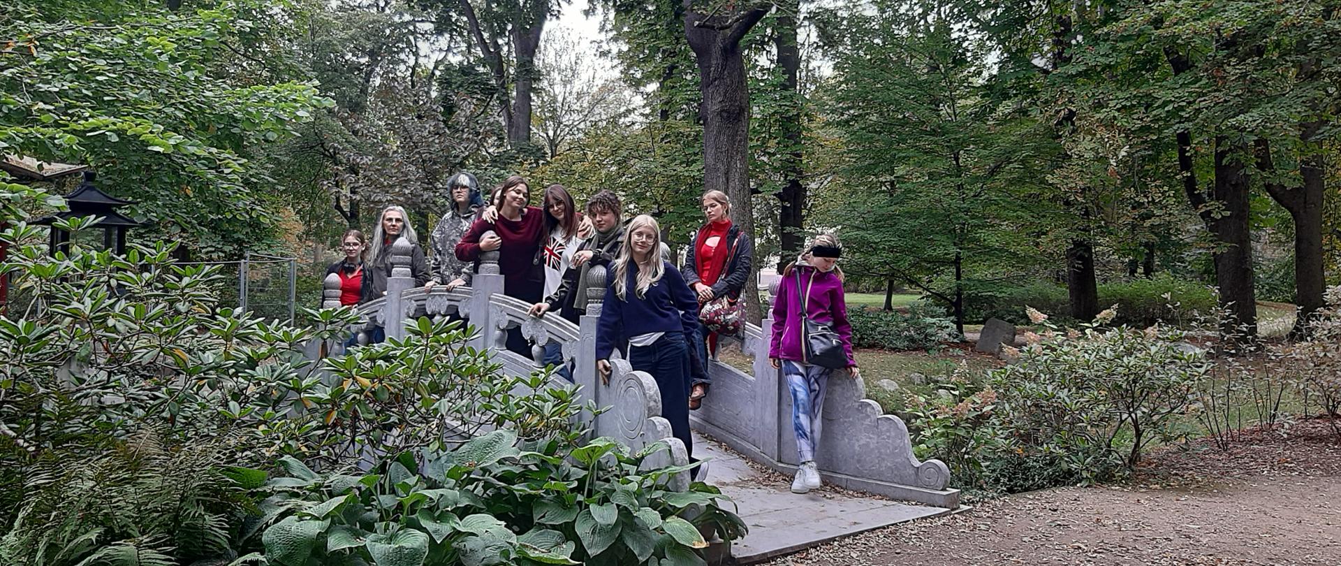 Grupa dziesięciu dziewcząt pozuje do zdjęcia stojąc na niewielkim rzeźbionym mostku wśród bujnej parkowej zieleni
