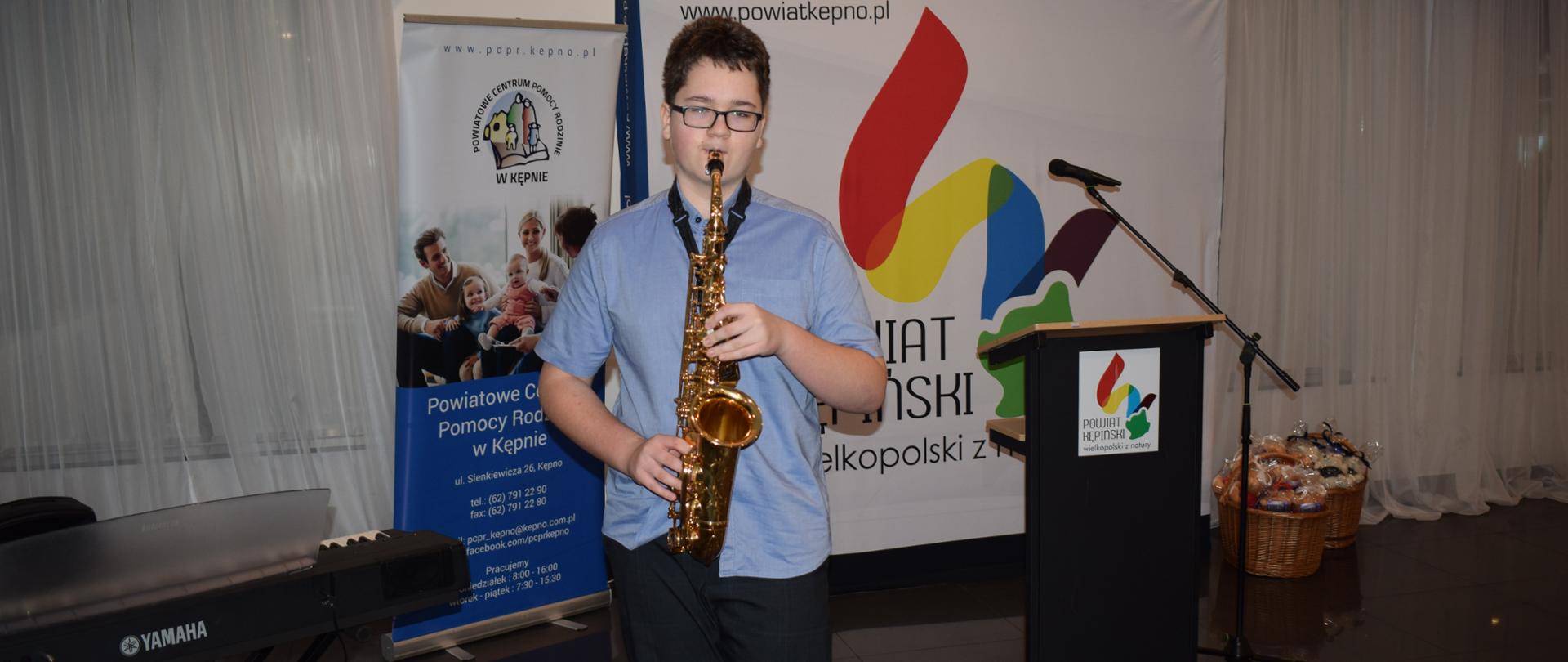 Na zdjęciu w centralnym miejscu znajduje się uczeń w niebieskiej koszuli grający na saksofonie. W tle widoczne są okna zasłonięte firanami, ponadto od lewej stoi instrument klawiszowy na krzyżowym statywie, obok (za uczniem) roll-up Powiatowego Centrum Pomocy Rodzinie i baner starostwa kępińskiego, następnie mównica konferencyjna z mikrofonem i wiklinowe kosze z maskotkami. 