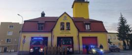 Siedmiu strażaków w piaskowych mundurach stojących na tle żółtej remizy Państwowej Straży Pożarnej z wieżą. Po lewej stronie widać wóz strażacki, a po prawej oznakowany radiowóz policyjny z włączonymi niebieskimi sygnałami świetlnymi oraz funkcjonariuszy policji.