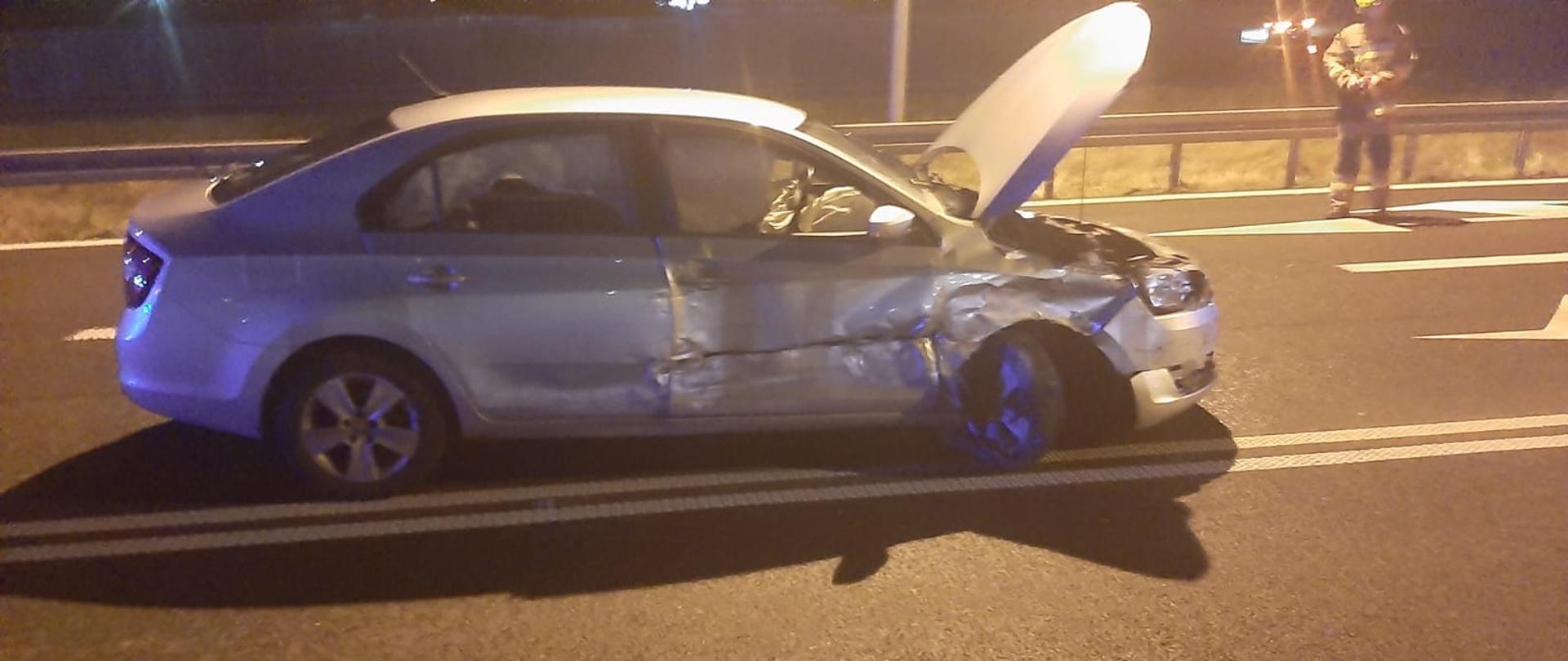 Srebrny samochód jest na drodze, uszkodzony przód i prawy bok, podniesiona maska, jest ciemno