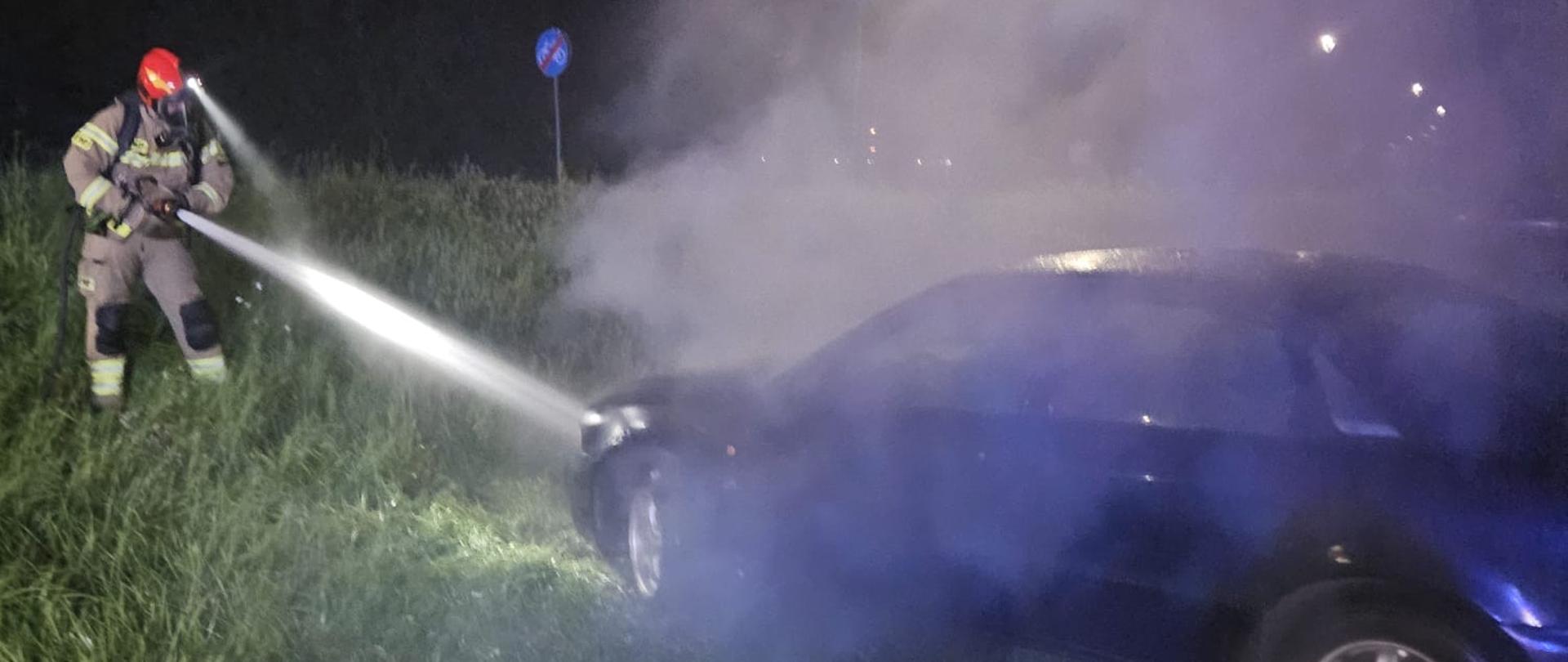 Zdjęcie przedstawia nocną akcję gaśniczą prowadzoną przez strażaka. Widać, że pali się samochód osobowy – z pojazdu wydobywa się gęsty dym, który unosi się w powietrzu. Strażak w pełnym umundurowaniu i czerwonym hełmie kieruje silny strumień wody z węża gaśniczego w stronę płonącego auta.
Teren jest porośnięty trawą, co sugeruje, że pojazd znajduje się poza drogą –na poboczu. W tle widoczne są znaki drogowe i uliczne lampy, co wskazuje na to, że zdarzenie miało miejsce w pobliżu jezdni. Scena jest dynamiczna, a nocna pora oraz intensywny dym potęgują dramatyzm sytuacji.
