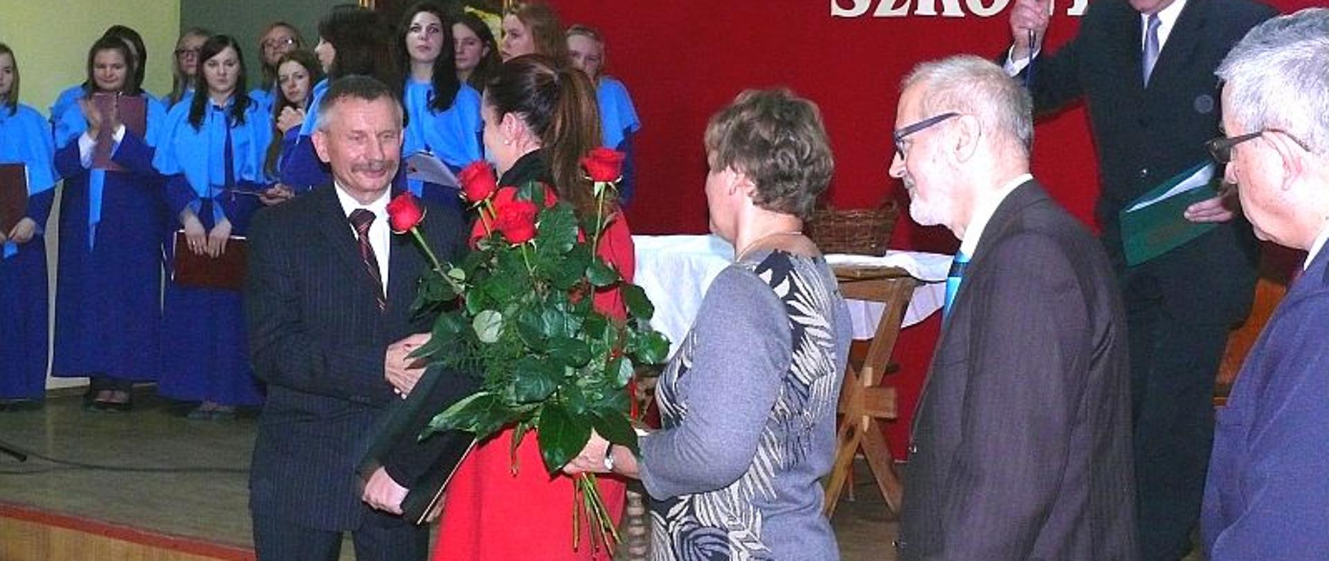 Dyrektor wręcza nauczycielą pamiątkowe dyplomy. W tle widać scenę a na niej chór.