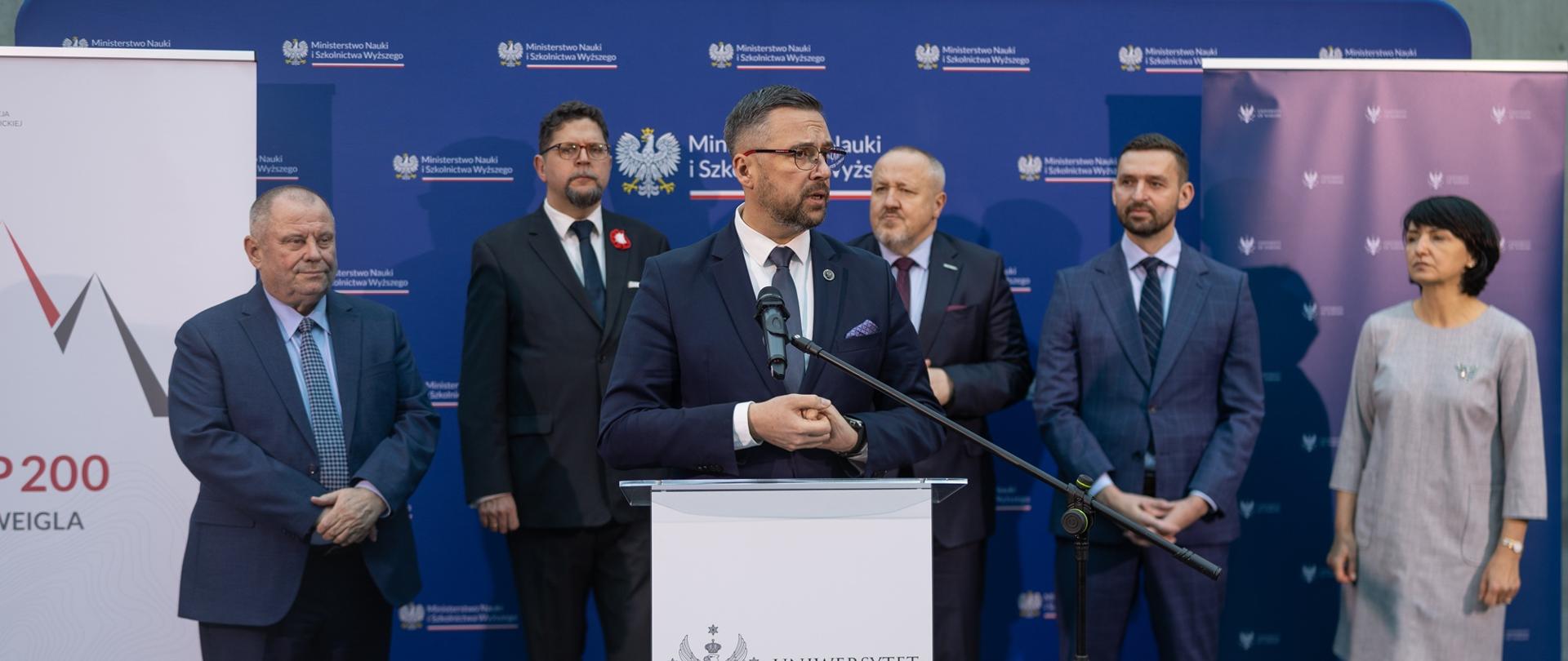 Za mównicą stoi minister Kulasek i mówi do mikrofonu, za nim przed granatową ścianką z napisem Ministerstwo Nauki i Szkolnictwa Wyższego stoi pięć osób - trzech mężczyzn w garniturach, wiceminister Szeptycki i kobieta w szarej sukience.