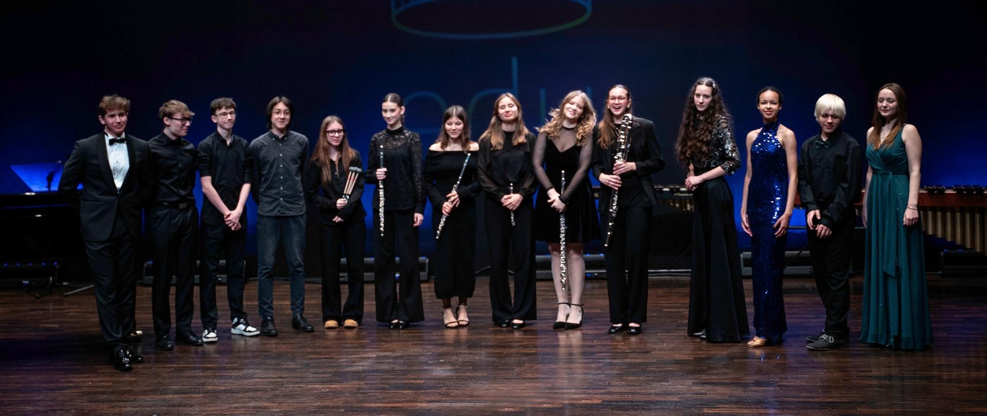 zdjęcie przedstawia wszystkich uczestników koncertu we wspólnym zdjęciu z instrumentami na scenie sali koncertowej