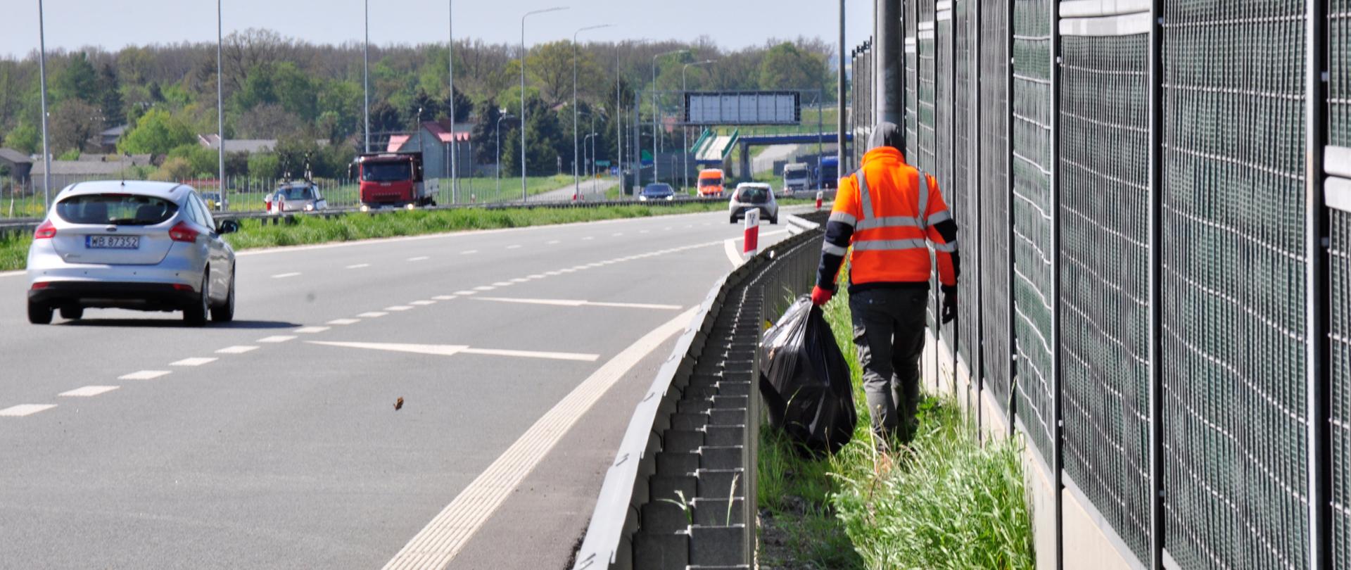 Zdjęcie przedstawia osobę ubraną w pomarańczową kamizelkę odblaskową, która porusza się wzdłuż pobocza autostrady, zbierając śmieci. Osoba ta trzyma duży czarny worek na śmieci i idzie obok metalowego ogrodzenia dźwiękochłonnego. Na drodze w tle widoczne są pojazdy, w tym srebrny samochód osobowy oraz czerwona ciężarówka. Po lewej stronie zdjęcia znajduje się pas zieleni oddzielający jezdnię od pobocza. W tle widoczne są drzewa oraz budynki, a dzień jest pogodny, z lekkim zachmurzeniem.