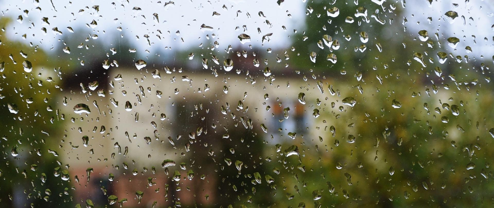 Na zdjęciu widoczne są krople deszczu na szybie. Daleko w tle budynek mieszkalny. 