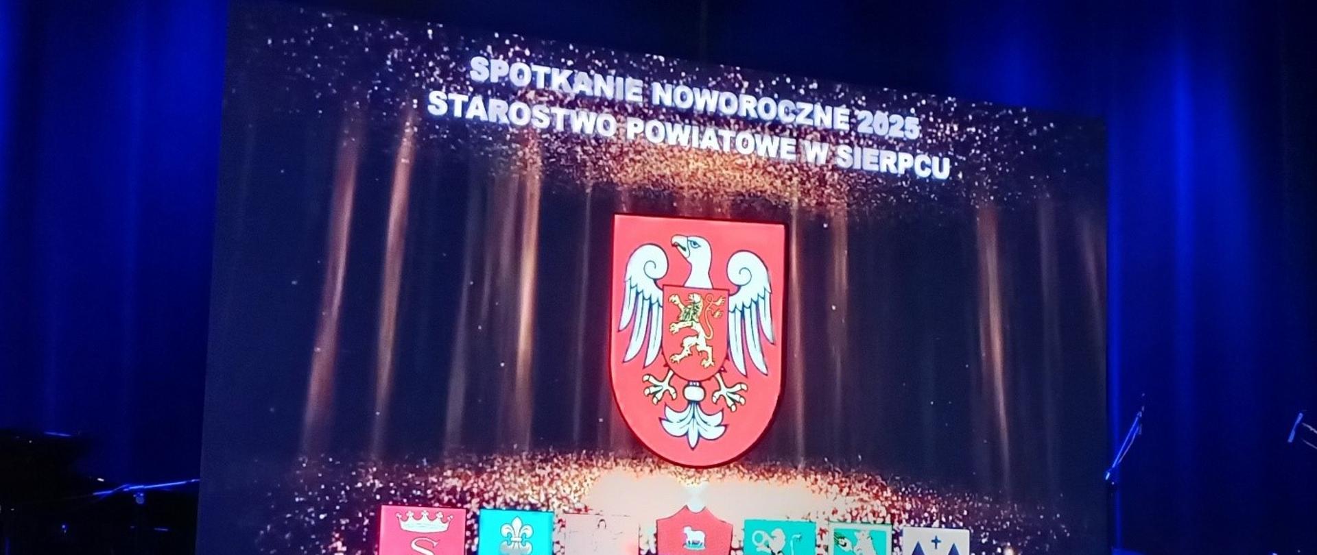 Zdjęcie przedstawia scenę oświetloną niebieskim światłem, na której znajduje się duży ekran z wyświetlonym napisem „SPOTKANIE NOWOROCZNE 2025 STAROSTWO POWIATOWE W SIERPCU”. Na ekranie widnieje herb powiatu sierpeckiego – biały orzeł z złotą koroną i lwem na czerwonej tarczy. Pod głównym herbem znajdują się mniejsze herby różnych gmin lub miejscowości wchodzących w skład powiatu. W tle sceny widoczne są elementy muzyczne, takie jak perkusja oraz mikrofony, co może sugerować część artystyczną wydarzenia.
