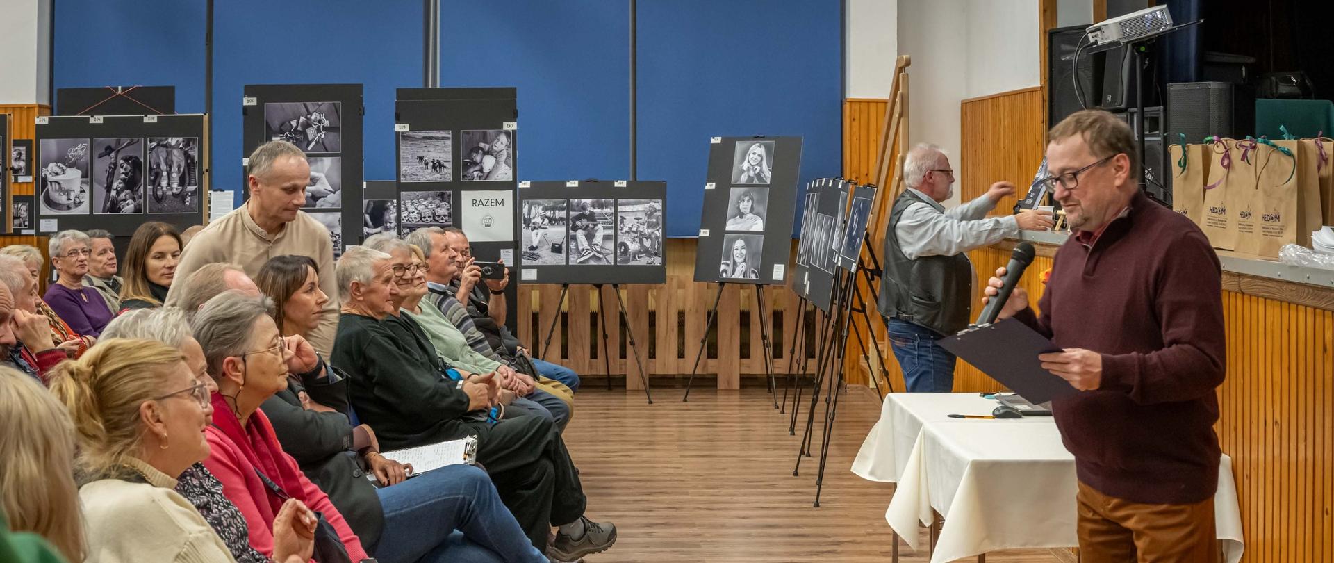Zdjęcie wykonane podczas wernisażu wystawy. Mężczyzna przemawia do zgromadzonej publiczności, która siedzi. W tle są czarno-białe zdjęcia wystawione na sztalugach. 