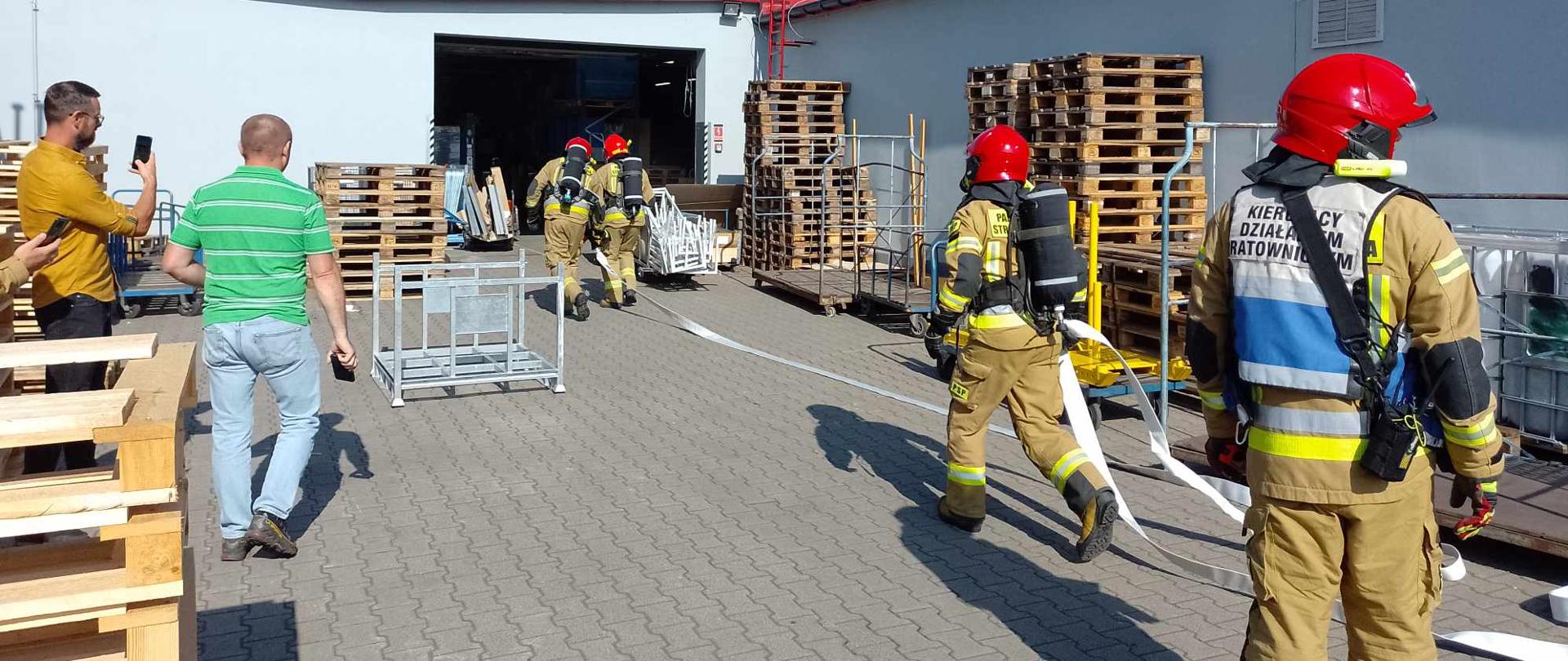 Trwają ćwiczenia w zakładzie produkcyjnym. Kierujący Działanaimi Ratowniczymi nadzoruje prace ratowników,. Wyznaczona rota w sprzęcie ochrony dróg oddechowych wchodzi do hali celem podjęcia działań gaśniczych. Rotawprowadza linie gasniczą