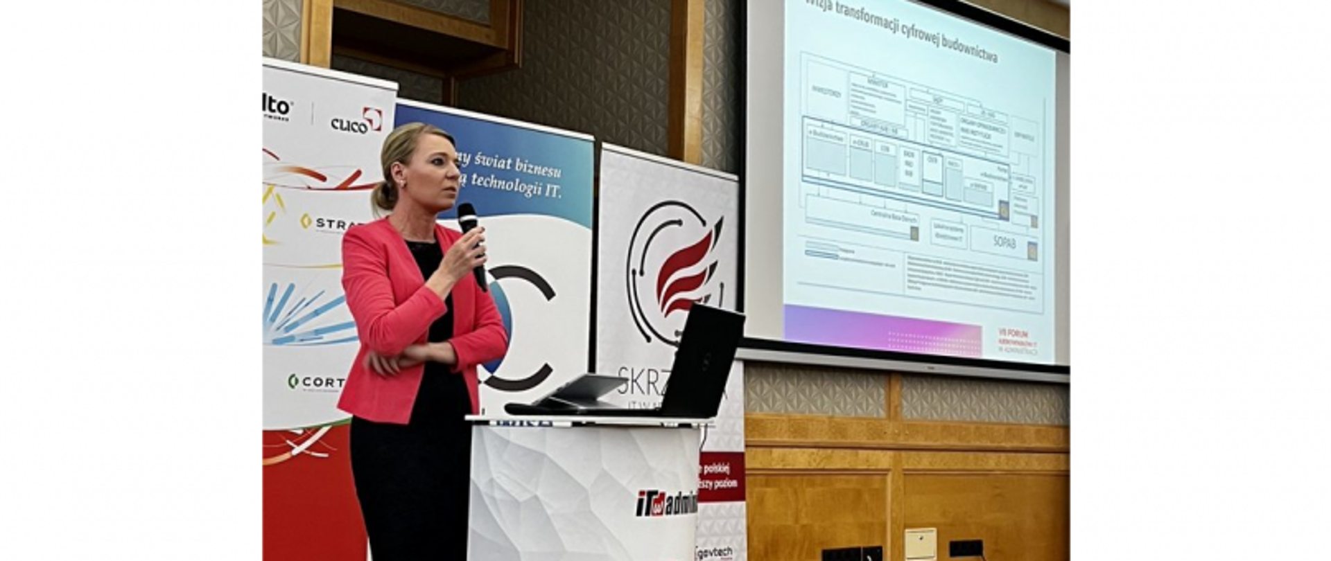 Osoba w czerwonej marynarce przemawia do mikrofonu przy mównicy z laptopem; w tle slajd „Mapa transformacji cyfrowej budownictwa” i banery konferencyjne.