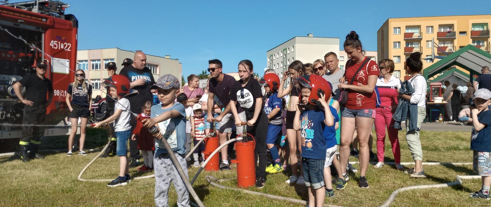 Chłopczyk trzymający prądownicę z wodą od hydronetki. Za nim wóz strażacki i tłum ludzi obecnych na pikniku. 