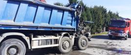Na zdjęciu widoczny jest uszkodzony samochód ciężarowy blokujący przejazd drogą powiatową. Z prawej strony częściowo jest widoczny samochód strażacki. W tle zabudowania miejscowości i roślinność przy drodze.