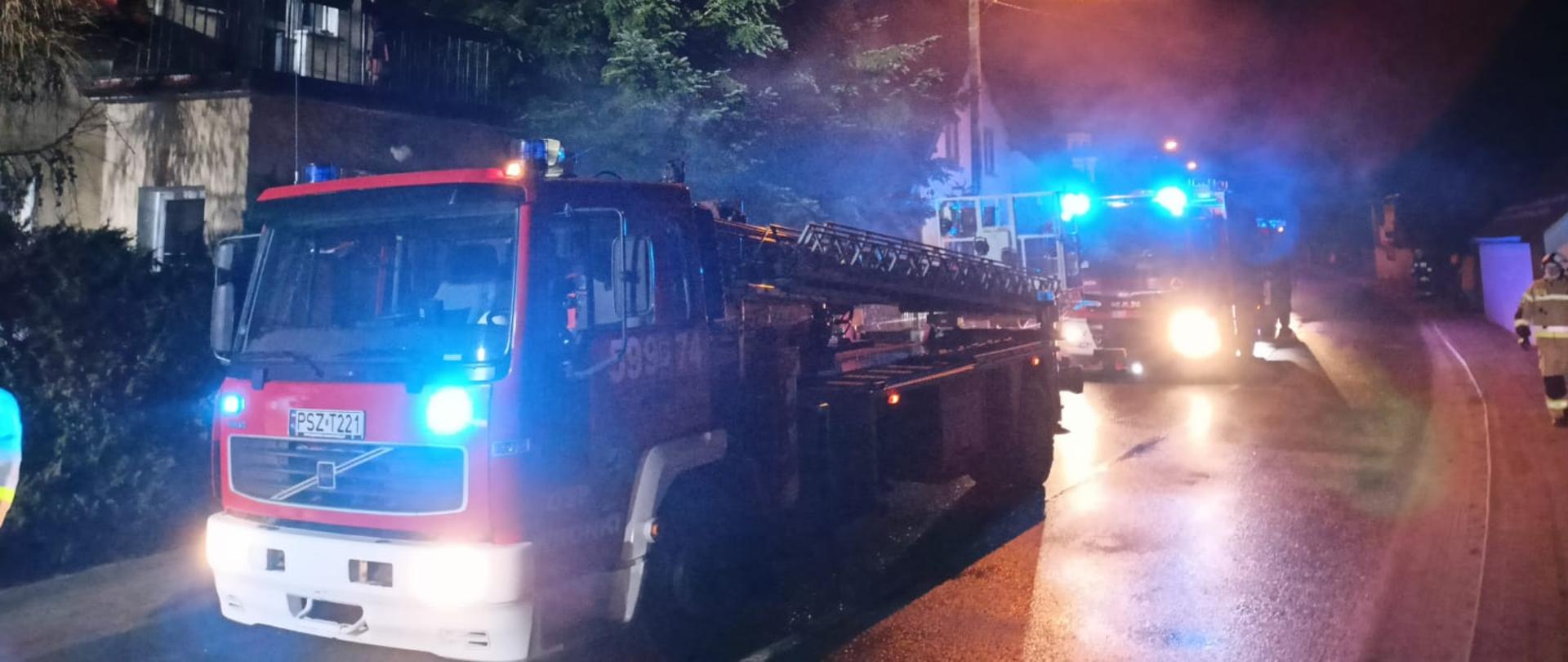 Widać budynek mieszkalny i samochód straży pożarnej, jest ciemno
