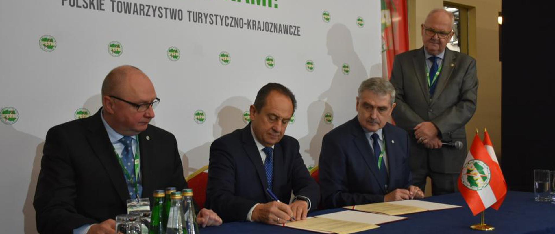 Zdjęcie przedstawia moment podpisania listu intencyjnego o współpracy Ministerstwa Sportu i Turystyki z Polskim Towarzystwem Turystyczno-Krajoznawczym. Na zdjęciu znajduje się czterech elegancko ubranych mężczyzn. Trzech z nich siedzi za stołem prezydialnym, jeden stoi tuż obok niego jako moderator uroczystości. W tle biała ścianka wizerunkowa PTTK z opisami i logotypami organizacji. 