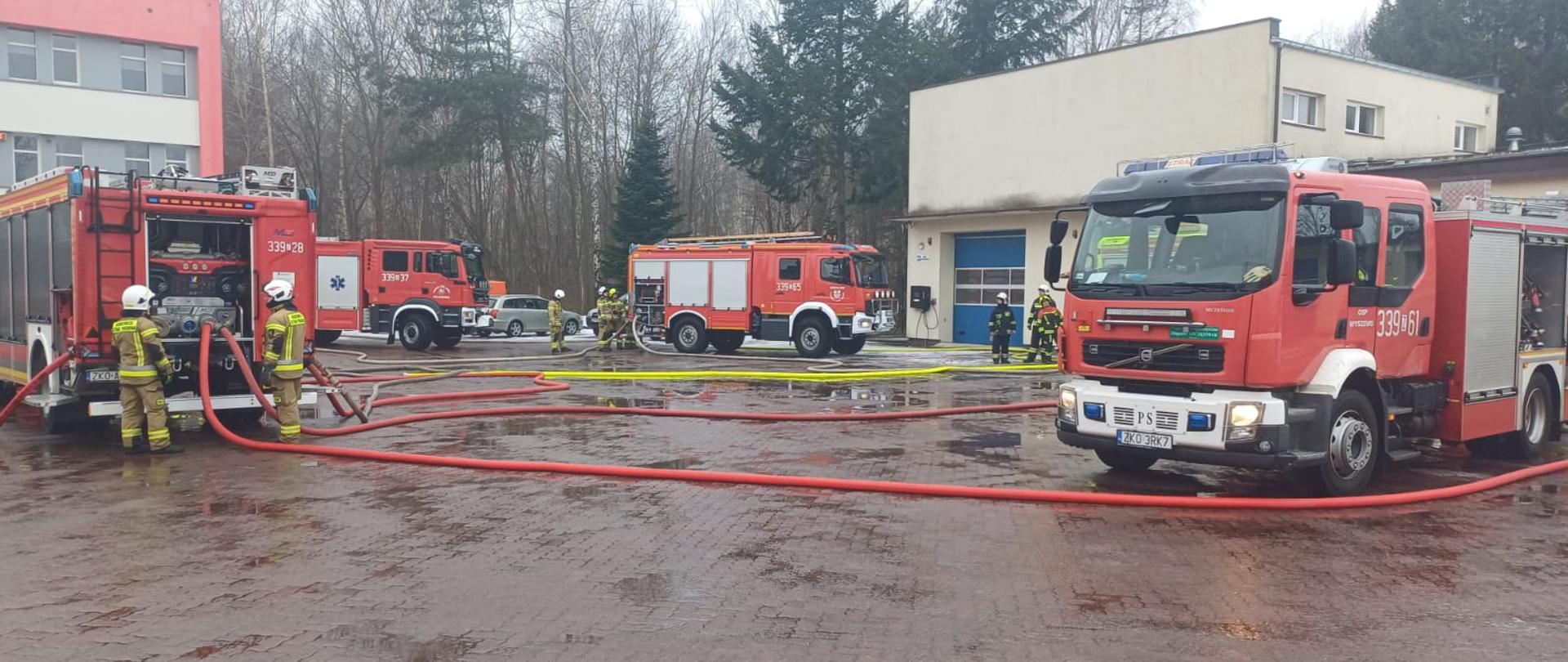 Samochody pożarnicze oraz strażacy podczas ćwiczeń z wykorzystaniem węży i sprzętu wodnego na placu PSP.