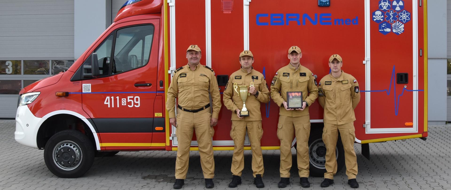 Zdjęcie przedstawia czterech dorosłych mężczyzn stojących na zewnątrz przed dużym, czerwono-białym pojazdem ratunkowym, który jest zaparkowany przed budynkiem.
Wszyscy mężczyźni są ubrani w jednolite, jasnobrązowe (piaskowe) mundury z czapkami z daszkiem w tym samym kolorze. Mundury mają pagony oraz emblematy.
Pojazd za nimi to samochód ratownictwa medycznego o intensywnie czerwonym nadwoziu z białym pasem na dole kabiny. 