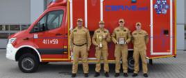 Zdjęcie przedstawia czterech dorosłych mężczyzn stojących na zewnątrz przed dużym, czerwono-białym pojazdem ratunkowym, który jest zaparkowany przed budynkiem.
Wszyscy mężczyźni są ubrani w jednolite, jasnobrązowe (piaskowe) mundury z czapkami z daszkiem w tym samym kolorze. Mundury mają pagony oraz emblematy.
Pojazd za nimi to samochód ratownictwa medycznego o intensywnie czerwonym nadwoziu z białym pasem na dole kabiny. 
