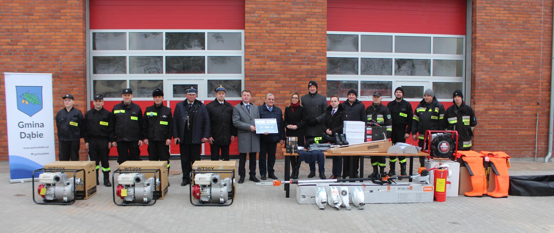 Zdjecie przedstawia uczestników przekazania sprzętu pożarniczego. Zdjęcie wykonano na tle remizy OSP Dąbie.