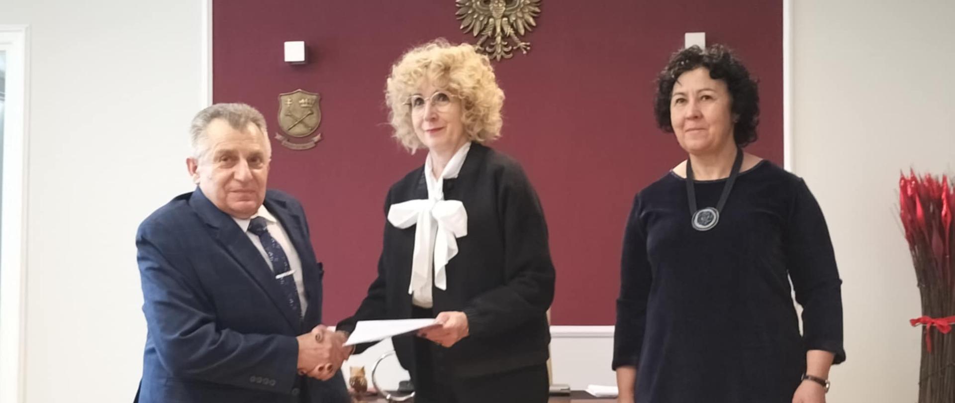 W centralnej części fotografii widzimy elegancko ubraną kobietę i mężczyznę w garniturze przekazujących sobie teczkę z dokumentem. Obok stoi druga elegancko ubrana kobieta, która jest świadkiem sytuacji. Wszystkie osoby są wyraźnie zadowolone i w dobrym nastroju. Cała scena odbywa się w gabinecie Rektora UJK w Kielcach. Wnętrze jest jasne, za plecami postaci widoczna jest bordowa tapeta z widocznym na niej godłem Państwa Polskiego. 