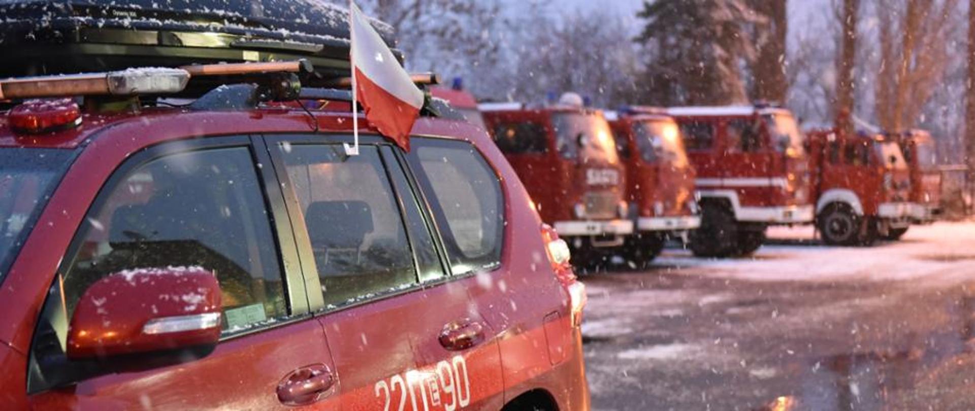 na zdjęciu bok samochody strażackiego osobowego w flaga Polski wpiętą w boczna szybę w tle samochody gaśnicze