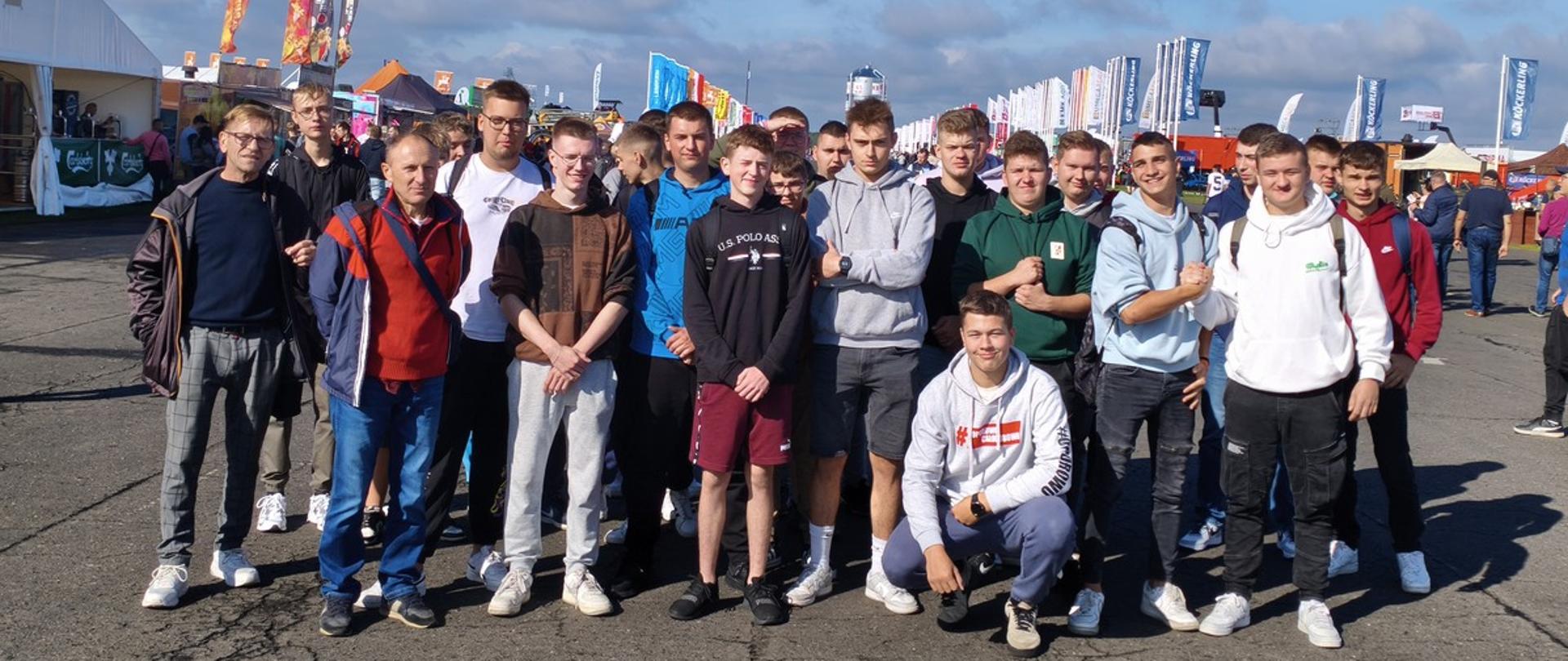 Grupa młodzieży wraz z opiekunami stoją przed wejściem na tergi Agro show Poznan. W tle banery reklamowe różnych firm oraz błękitne niebo.