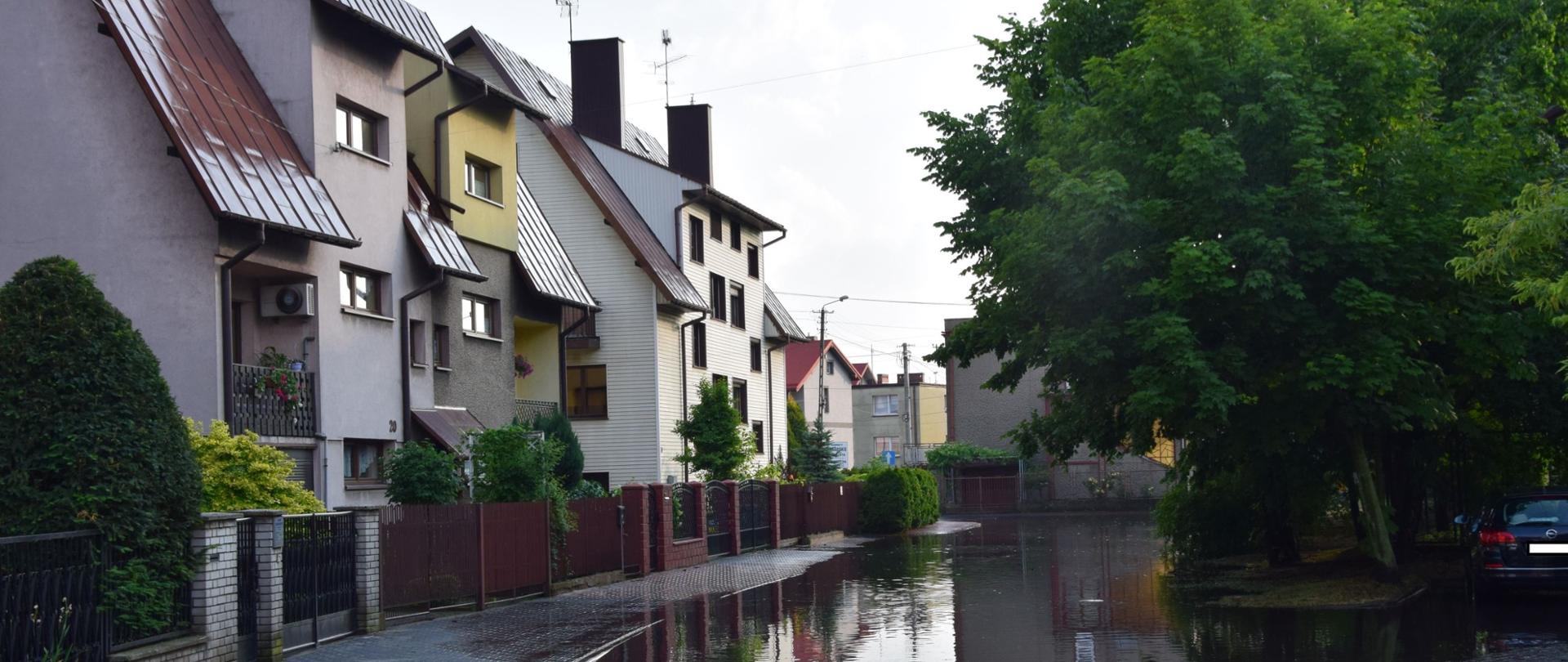 Zdjęcie przedstawia zalaną drogę na osiedlu, widoczna woda na całej szerokości jezdni oraz domy mieszkalne 